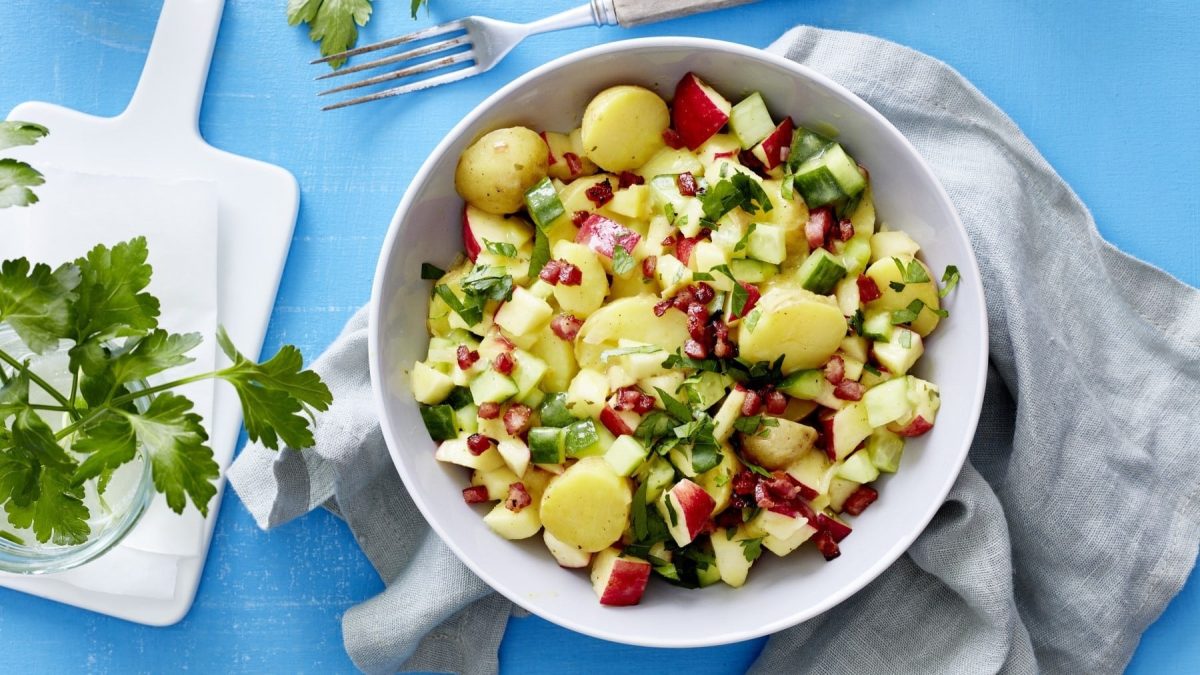 Kartoffel-Gurken-Salat mit Apfel, Speckwürfeln und frischer Petersilie, serviert in einer großen Salatschale auf einem blauen Küchentisch.