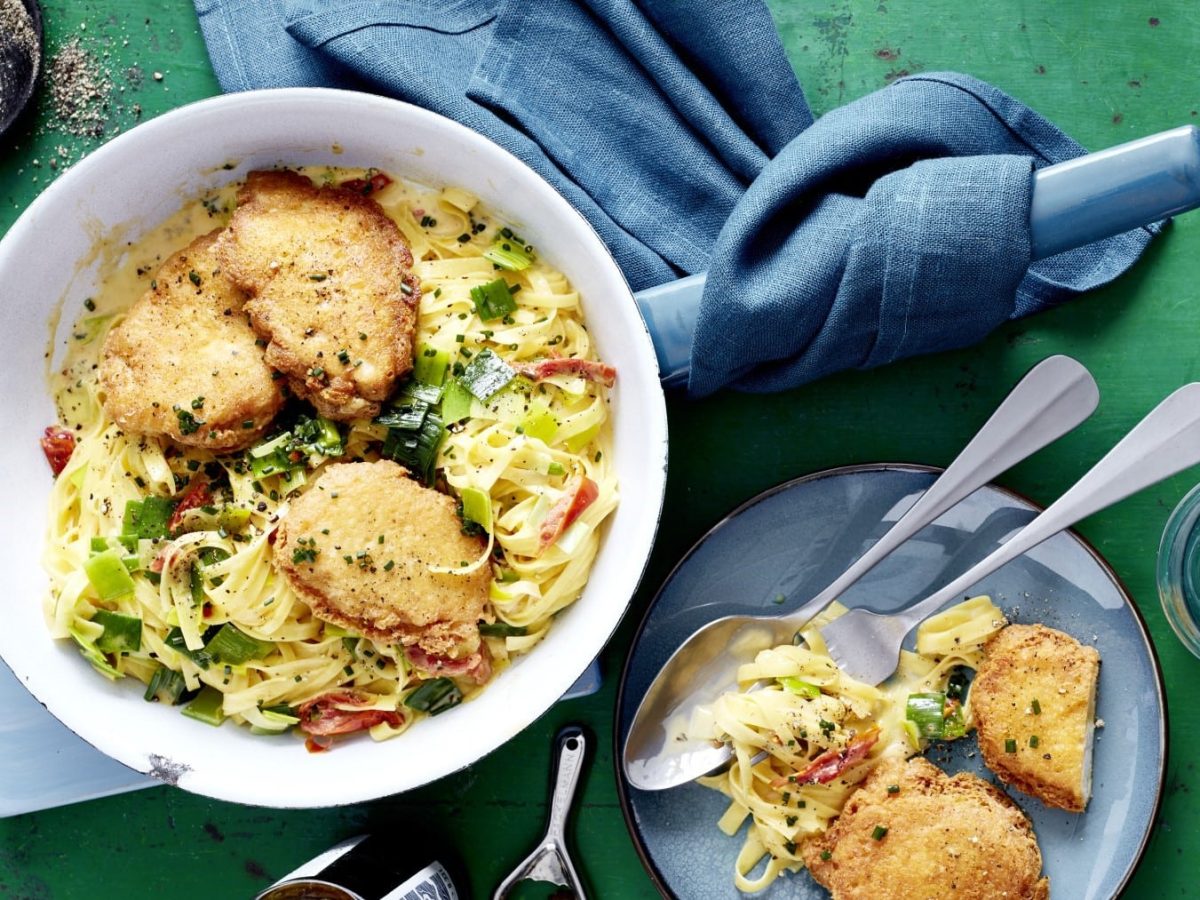 Parmesan-Schnitzel mit Porree-Carbonara, serviert in einer großen Schale und garniert mit Schnittlauch und grobem Pfeffer auf einem grünen Esstisch.