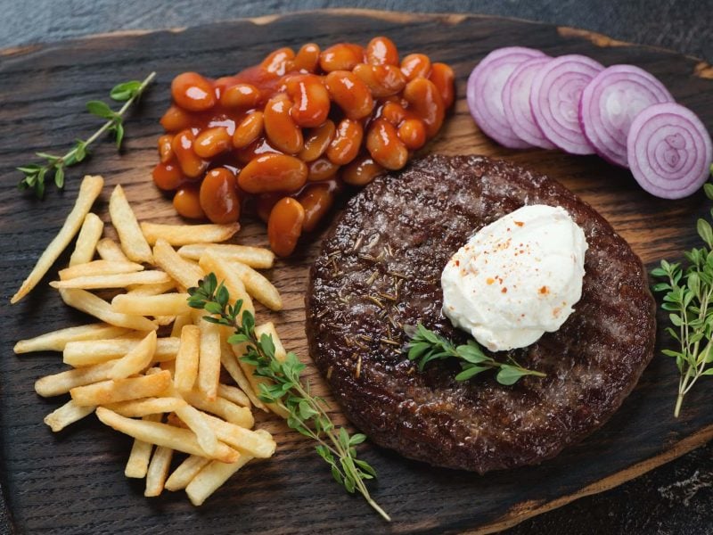 Pljeskavica mit Rahmkäse, Bohnen, Pommes, Zwiebeln und Kräutern auf Holzbrett. Draufsicht.