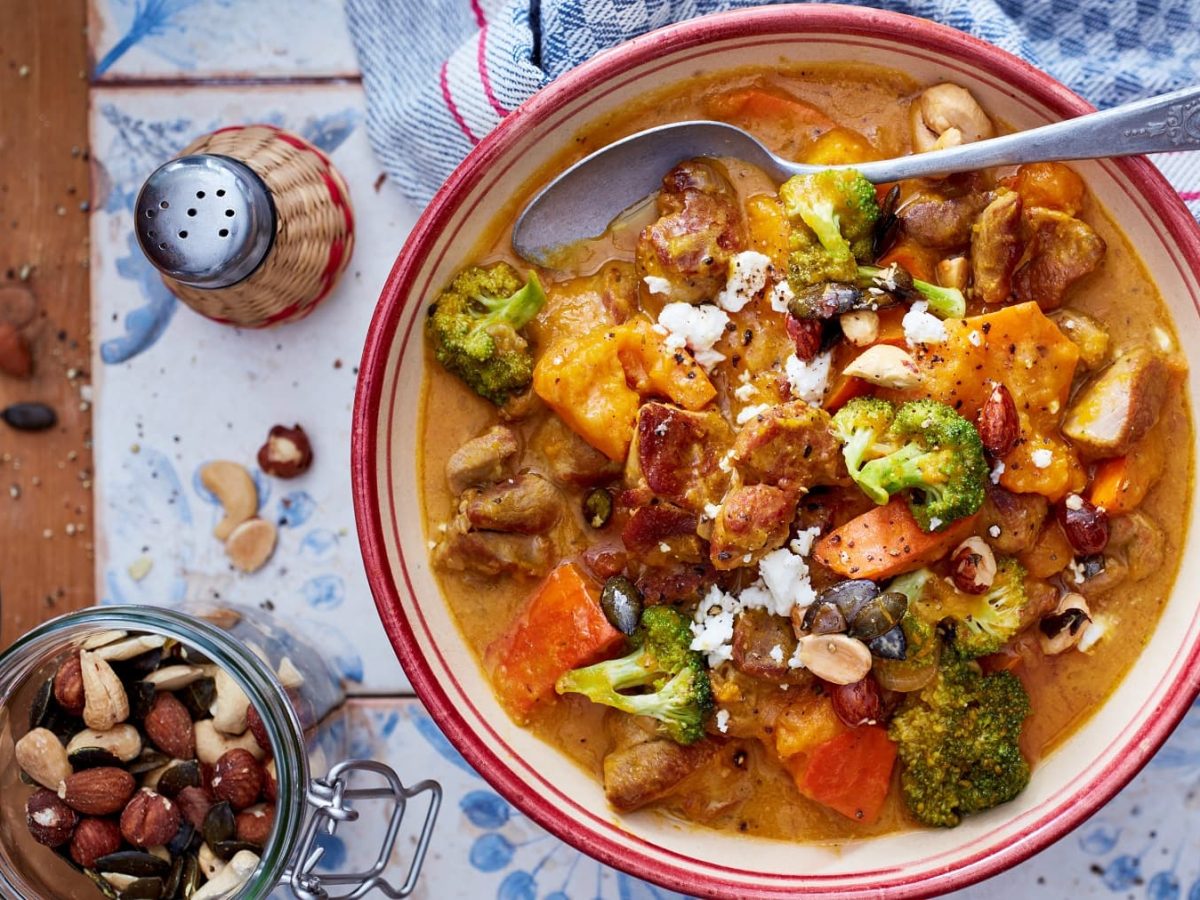 Putengulasch mit Kürbis und Honignüssen in einer Schüssel mit Löffel neben Salzstreuer und Nüssen vor hellem Hintergrund.