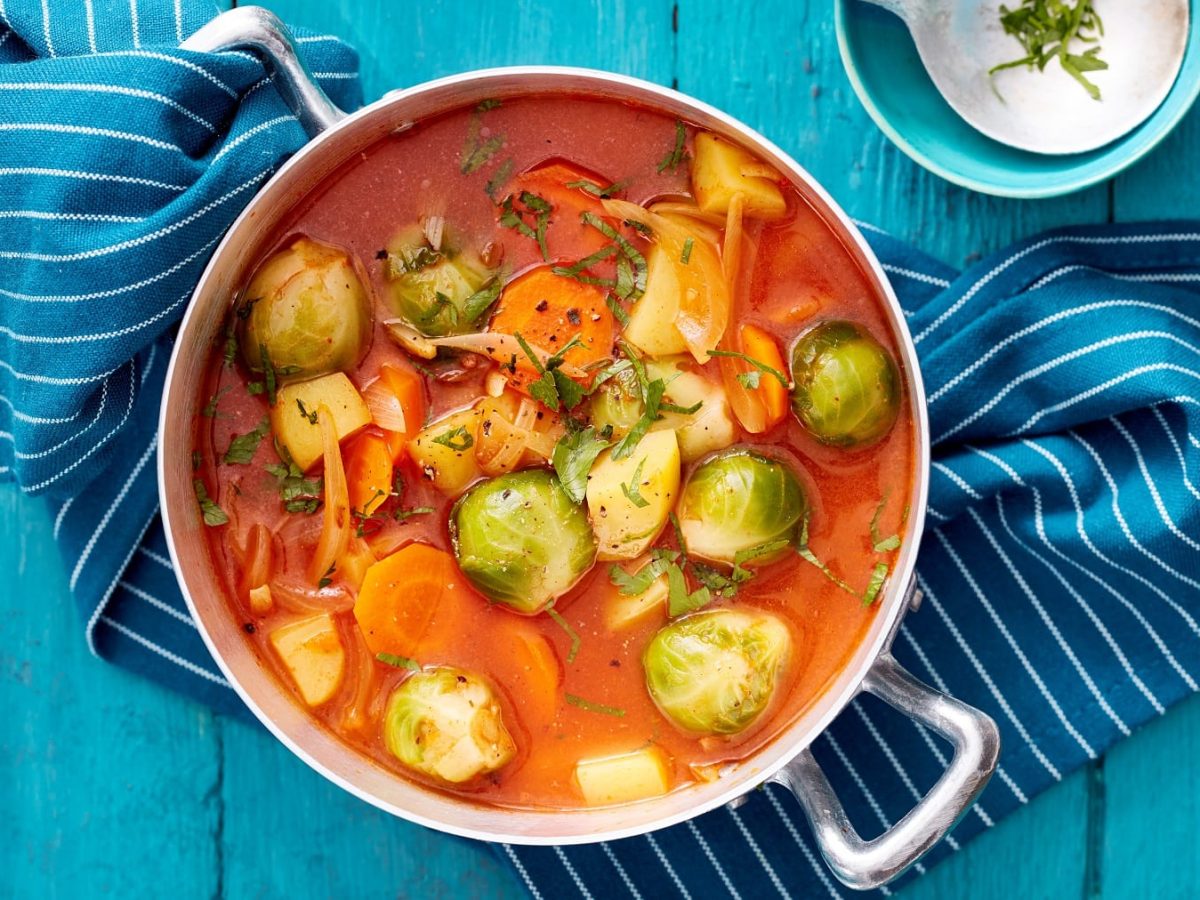 Rosenkohl-Kartoffel-Eintopf mit Möhren und frischen Kräutern, serviert im großen Topf auf einem blauen Tisch.