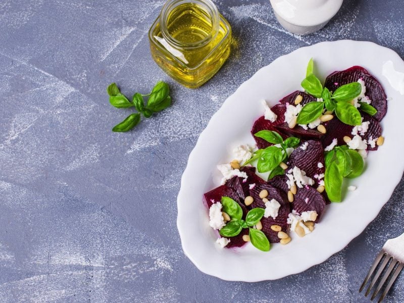 Rote-Bete-Carpaccio mit Feta auf blauem Untergrund, daneben Olivenöl und Pinienkerne