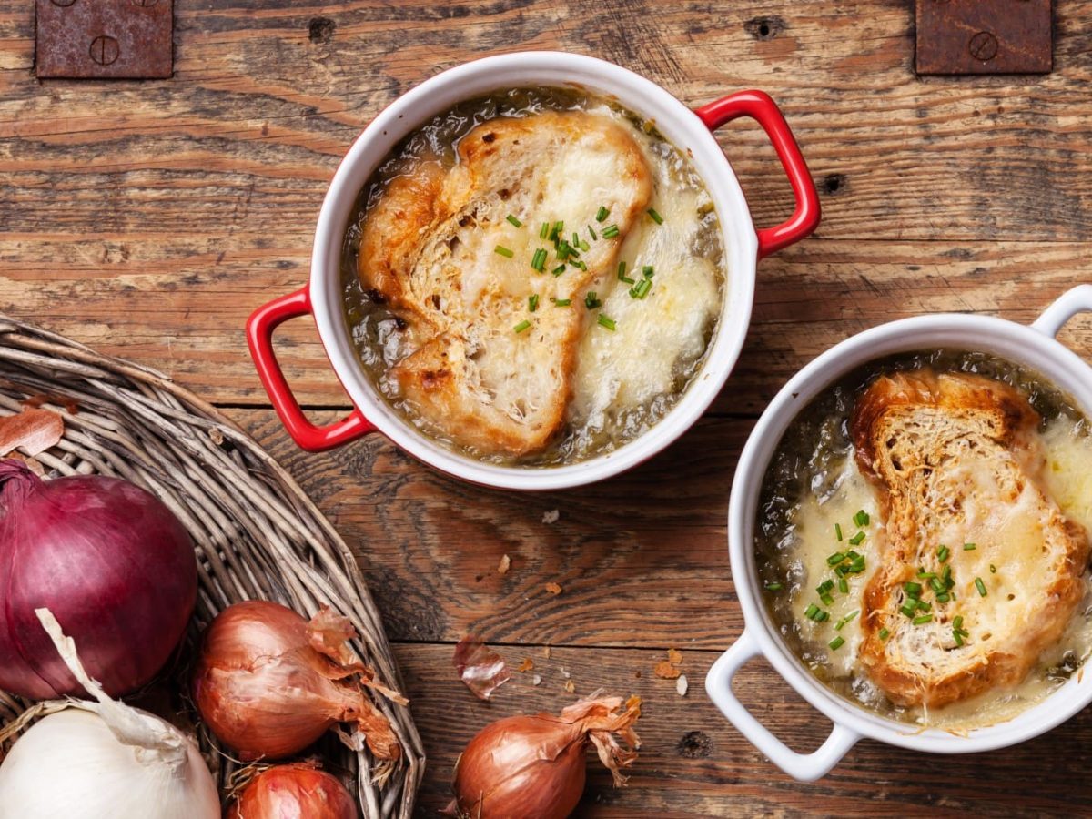 Soupe à l'ognion in zwei Schalen. Daneben ein Korb mit Zwiebeln und Knoblauch.