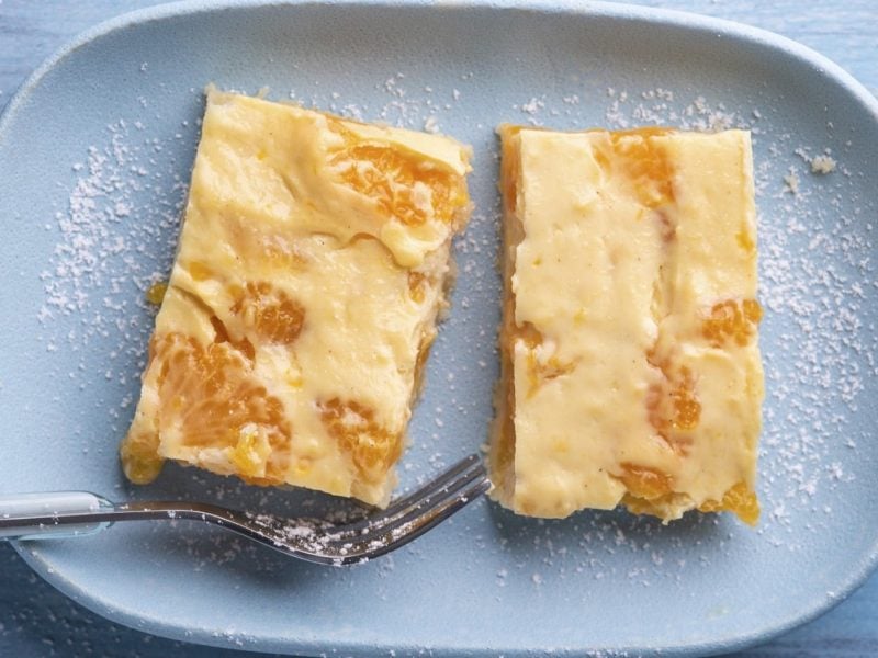 Auf einem Teller liegen zwei Stück veganer Mandarinen-Quark-Kuchen. Sie sind mit Puderzucker bestäubt.