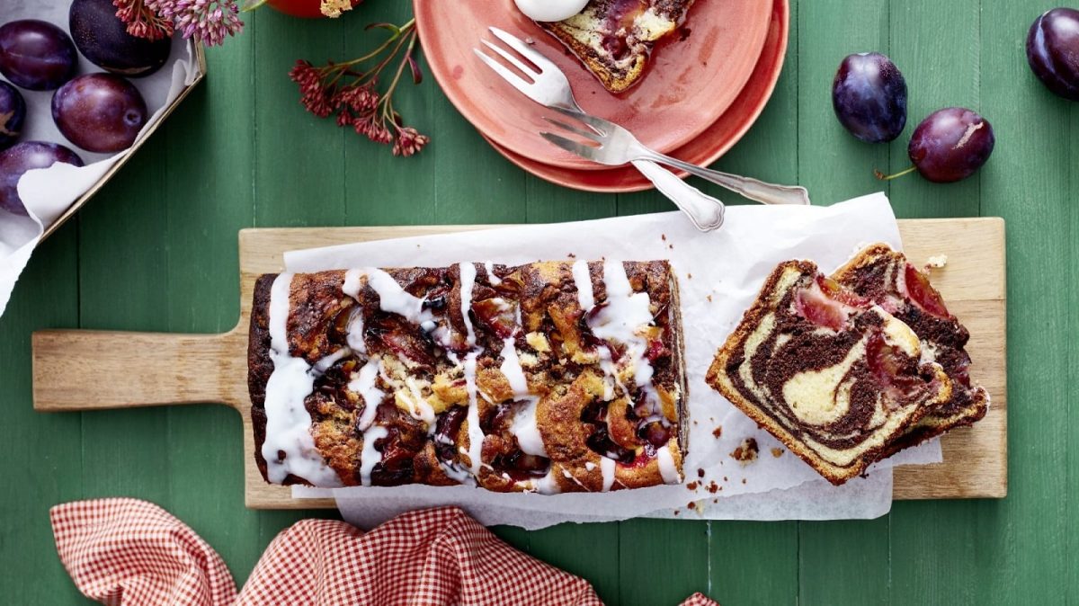 Auf einer Holzplatte liegt ein Zwetschgen Marmorkuchen. Er ist mit einem Puderzuckerguss überzogen.