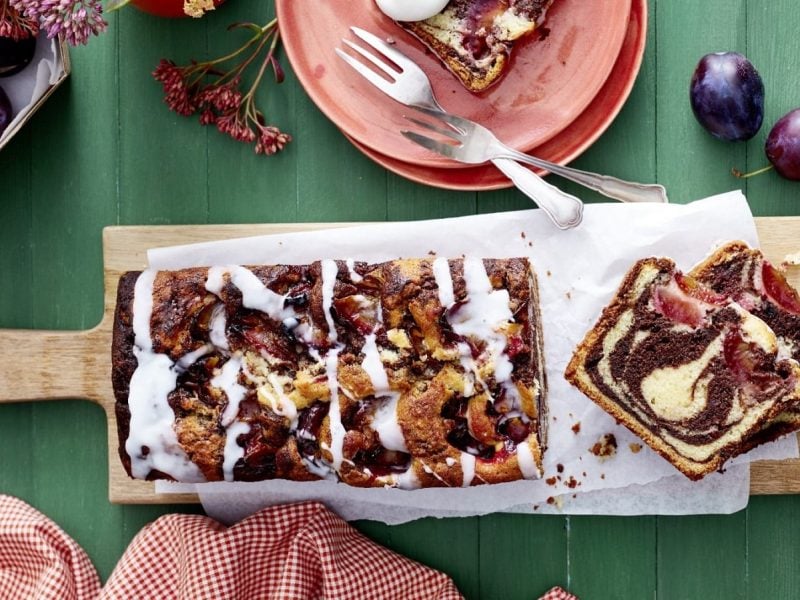 Auf einer Holzplatte liegt ein Zwetschgen Marmorkuchen. Er ist mit einem Puderzuckerguss überzogen.