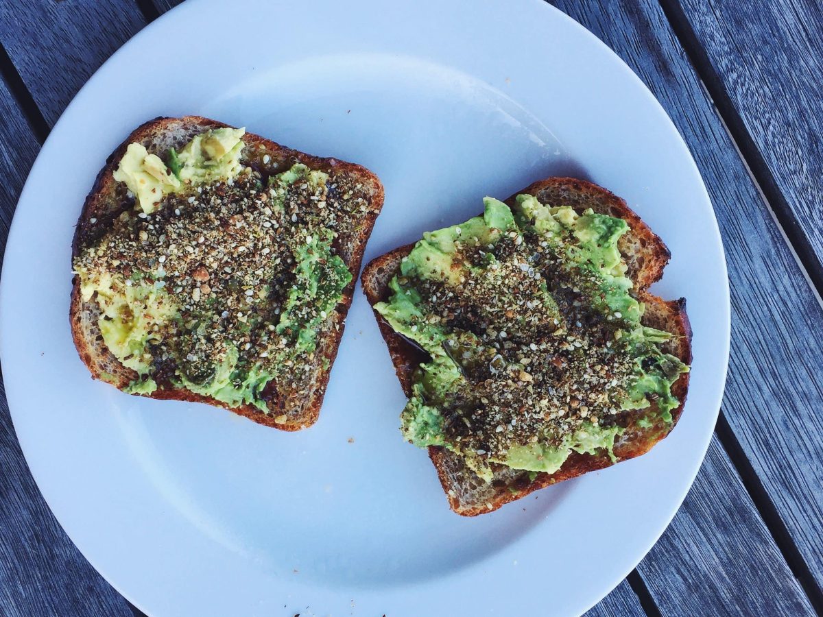 Zwei Toasts auf einem weißen Teller mit Avocado und Dukkah bestreut. Der Untergrund ist ein rustikaler Holztisch.