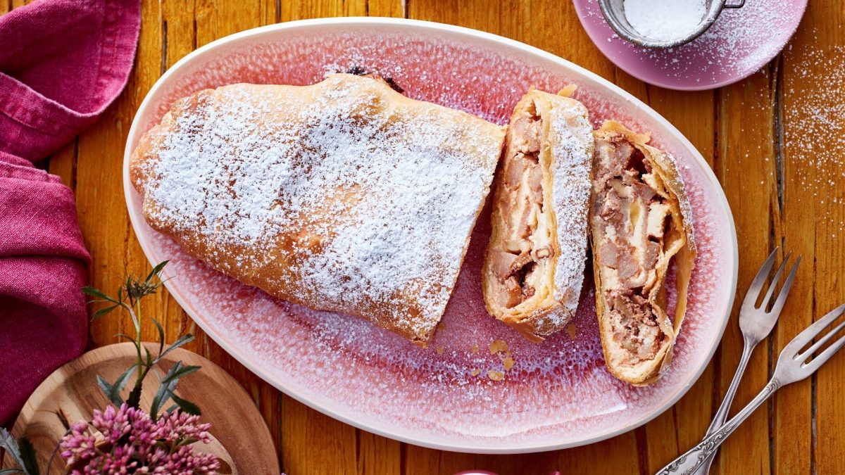 Angeschnittener Birnen-Käsekuchen-Strudel auf einem ovalen rosa Teller neben Besteck vor Holzhintergrund