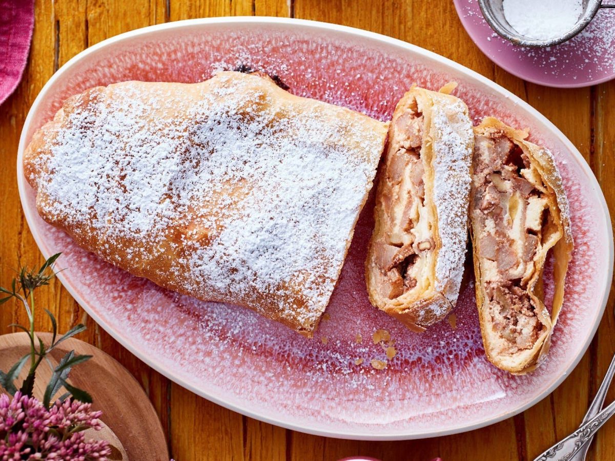 Angeschnittener Birnen-Käsekuchen-Strudel auf einem ovalen rosa Teller neben Besteck vor Holzhintergrund