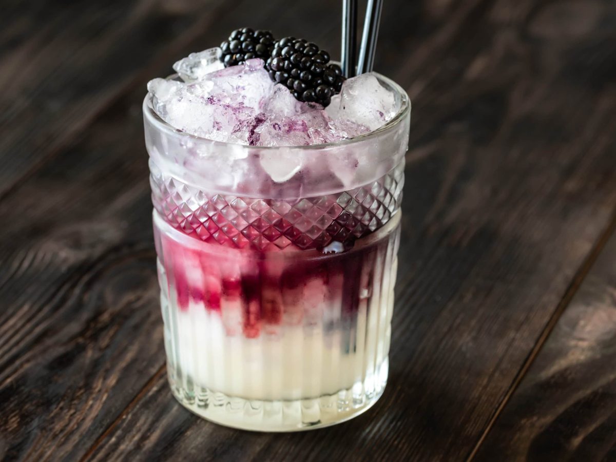 Bramble Gin-Cocktail mit Brombeeren in Tumbler mit frischen Brombeeren und Strohhalm auf dunklem Untergrund. Frontalaufnahme.