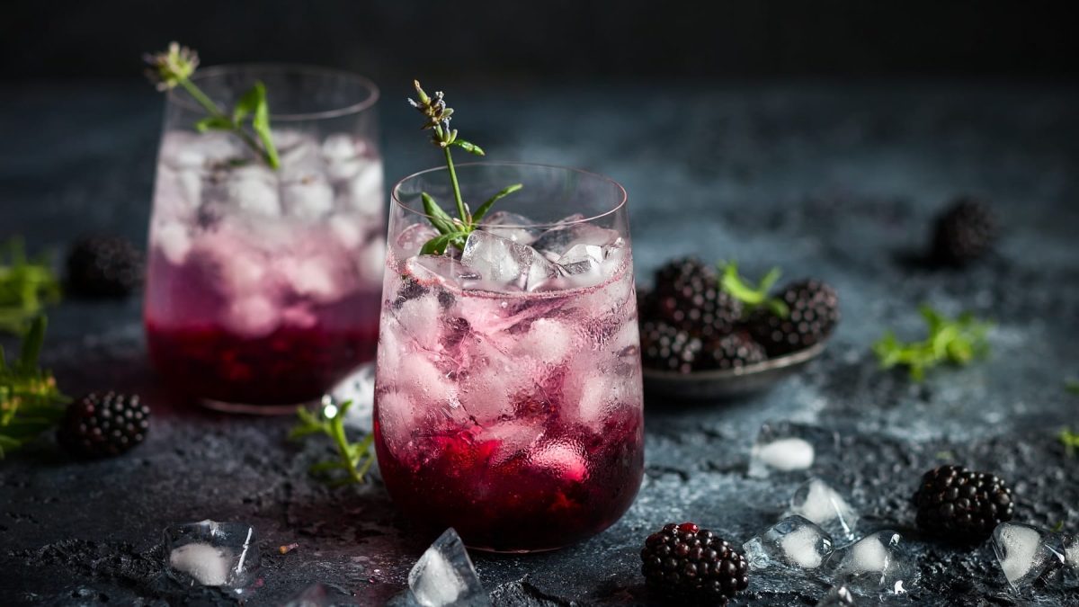 Zwei Gläser Blackberry Fizz mit Eiswürfeln, drumherum frische Brombeeren und Eiswürfel.