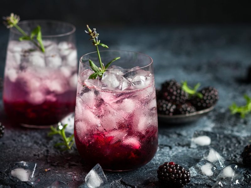 Zwei Gläser Blackberry Fizz mit Eiswürfeln, drumherum frische Brombeeren und Eiswürfel.