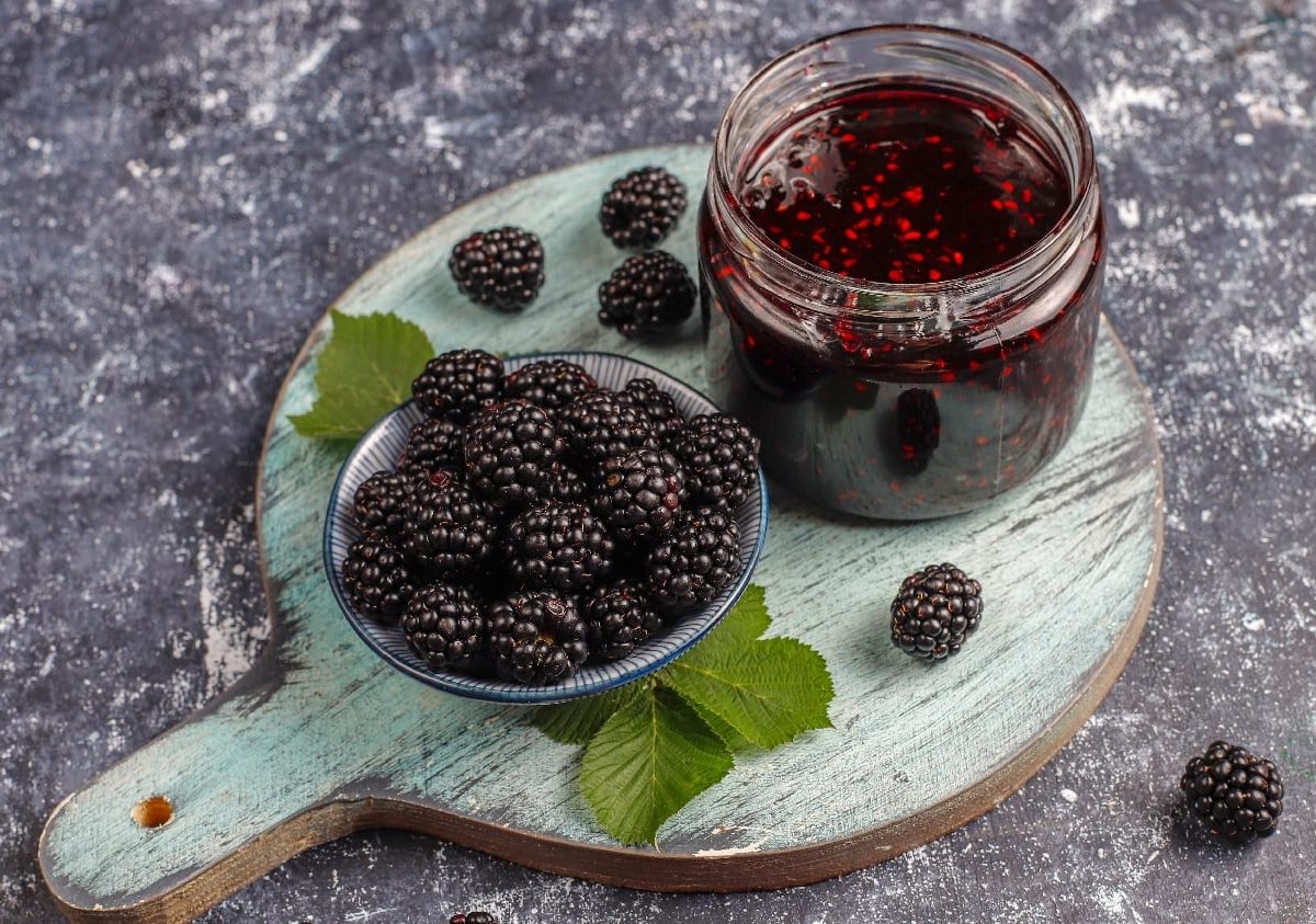 Ein Glas Brombeer-Marmelade auf einem Holzbrett mit frischen Brombeeren.