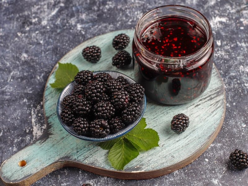 Ein Glas Brombeer-Marmelade auf einem Holzbrett mit frischen Brombeeren.