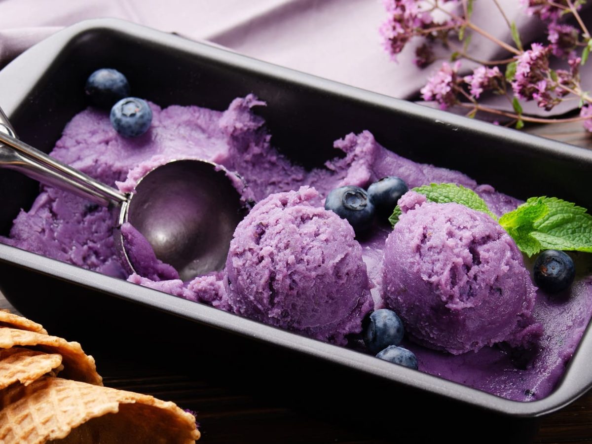 Eine Kastenform mit Brombeer-Nicecream, darauf Eiskugeln und ein Eisportionierer.