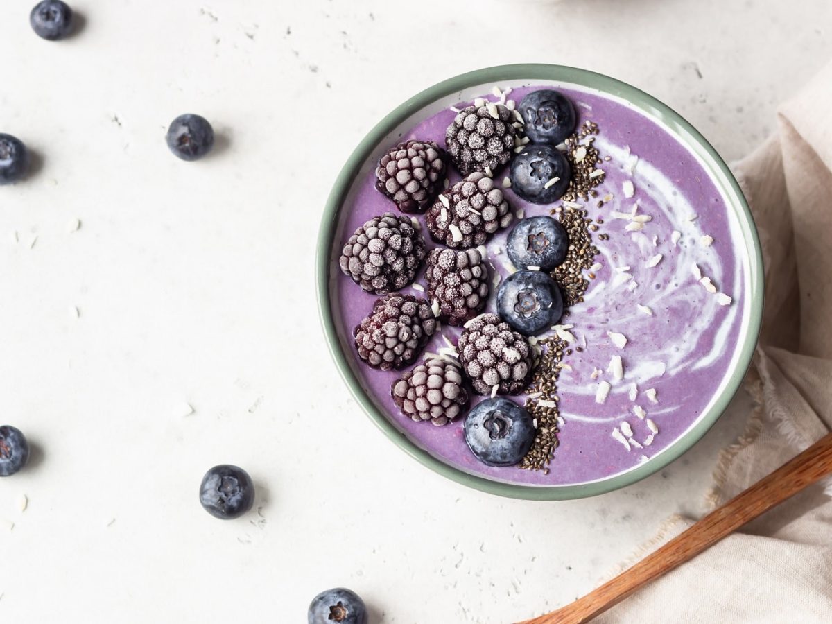 Eine Brombeer-Smoothie-Bowl mit Kokosflocken und gefrosteten Brom- und Heidelbeeren in der Draufsicht, daneben liegen ein paar Heidelbeeeren.