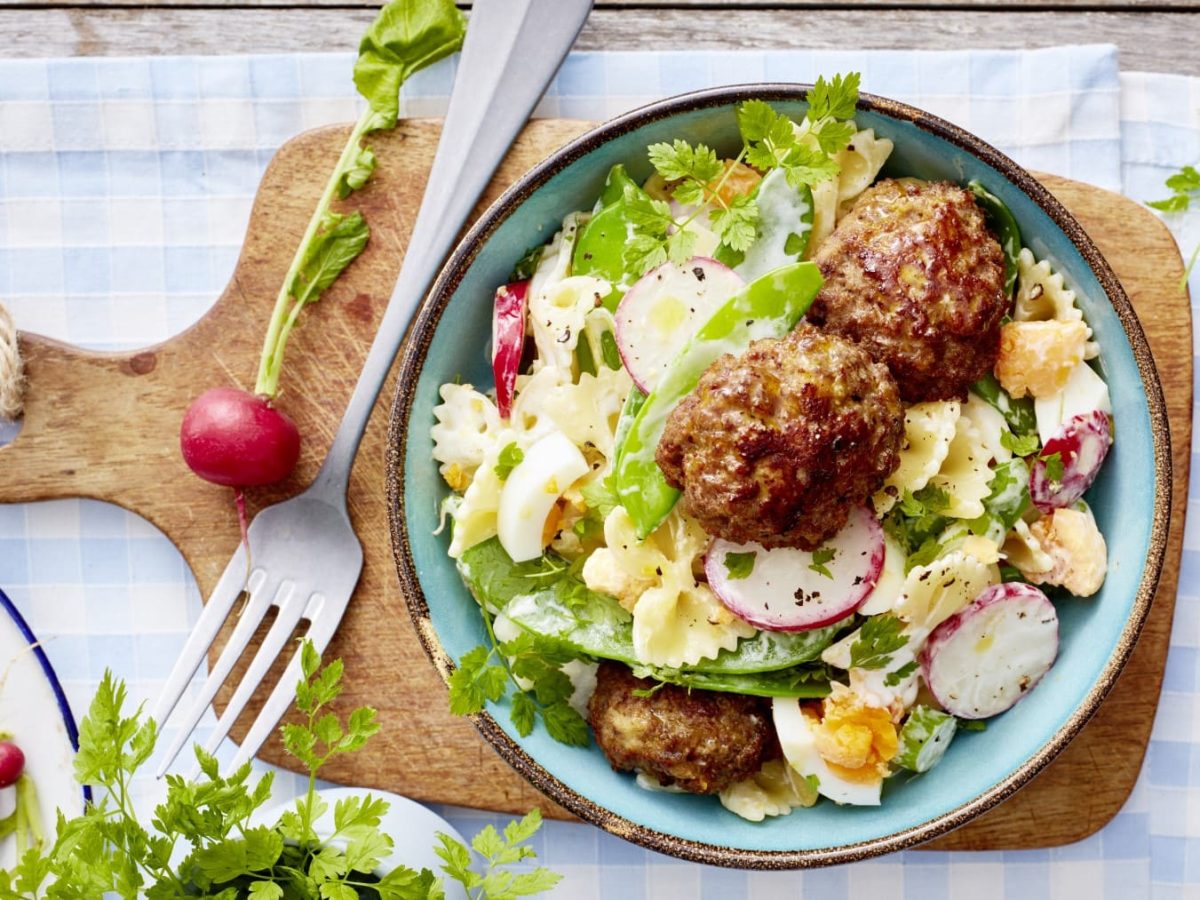 Buletten mit Nudelsalat in einem Teller auf einem Holzbrett neben Radieschen und einer Gabel.