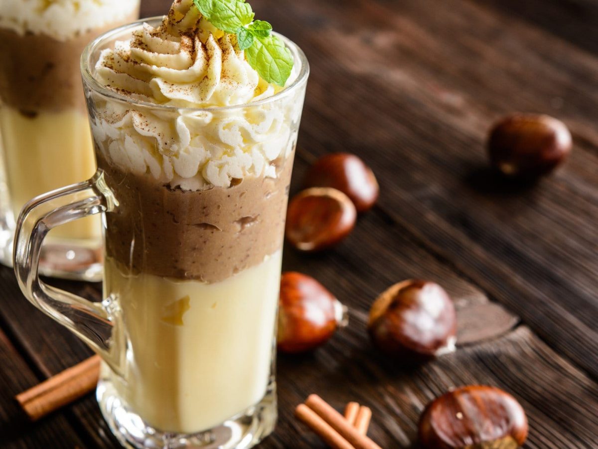 Cognac-Vanillepudding mit Maronenpüree und Pistazien-Sahne in Glas auf dunklem Holz. Daneben Maronen und Zimtstangen. Frontalansicht.