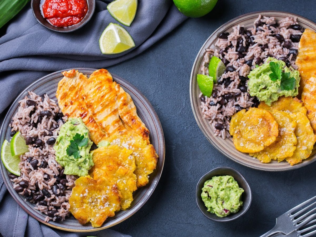 Zwei Teller mit Arroz Congrís con Tostones y Pollo neben kleinen Schalen mit Guacamole und Tomatensauce vor dunklem Hintergrund.