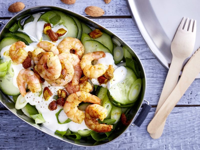 Curry-Garnelen mit Gurkensalat in einer Brotdose. Daneben liegt hölzernes Besteck.