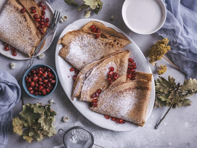 Einige Dinkel-Crêpes mit Granatapfelkernen auf Tellern neben herbstlicher Deko vor hellem Hintergrund.