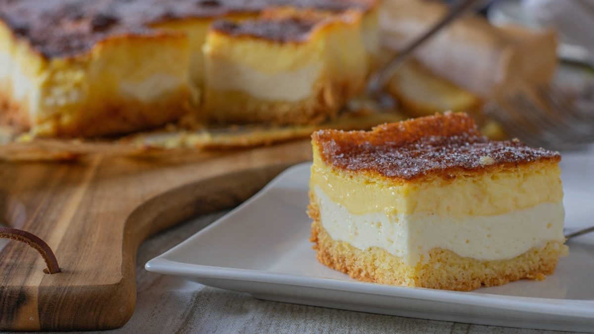 Ein Stück Eierschecke, bestehend aus einer Schicht Teig, einer Schicht Puddingcreme und einer Schicht Quarkcreme, serviert auf einem weißen Teller neben den übrigen Stücken im Hintergrund.