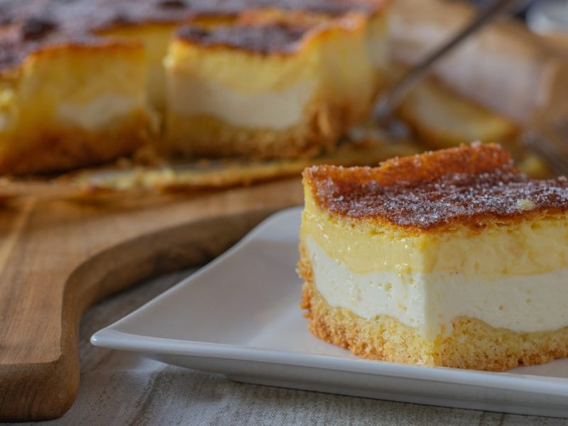 Ein Stück Eierschecke, bestehend aus einer Schicht Teig, einer Schicht Puddingcreme und einer Schicht Quarkcreme, serviert auf einem weißen Teller neben den übrigen Stücken im Hintergrund.