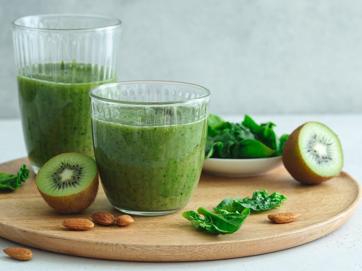 Zwei Gläser Spinat-Smoothie mit Kiwi auf einem Holzbrett neben frischen Früchten.