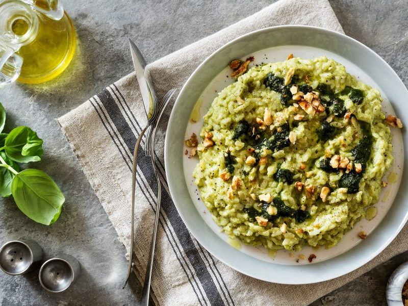 EIn Teller mit Zucchini-Risotto mit Pesto auf einem grauen Geschirrtuch neben frischen Zutaten.