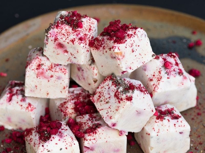 Mehrere Würfel Frozen Joghurt mit Himbeeren liegen auf einem dunklenTeller.