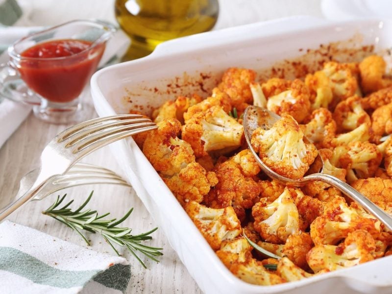 Eine Auflaufform gebackener Blumenkohl mit Tomatensauce, darin ein Löffel.