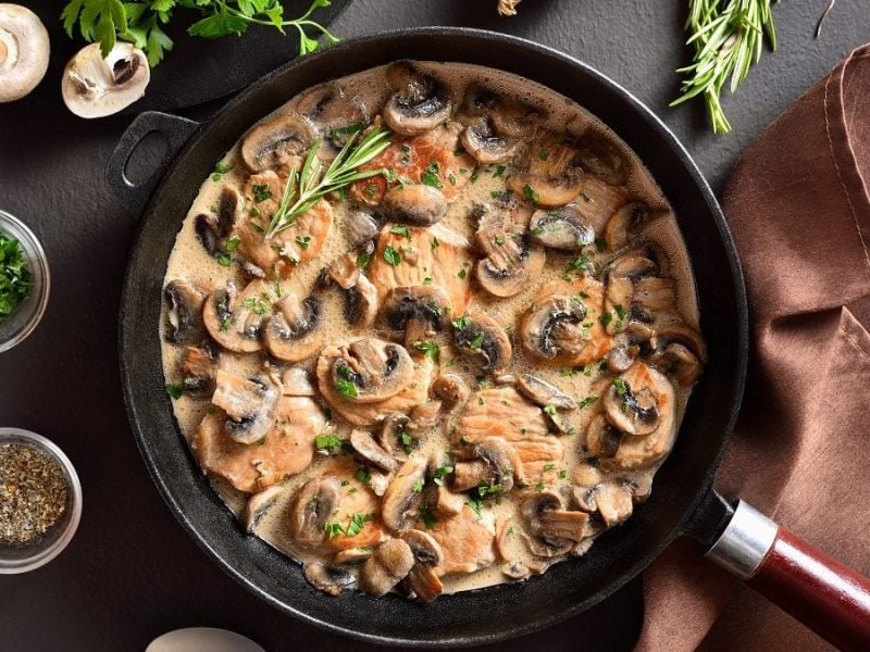 Draufsicht: Eine Pfanne mit einem Geflügel-Ragout steht auf einem dunklen Untergrung. Drum herum stehen und liegen Gläser mit Kräutern und Gewürzen.