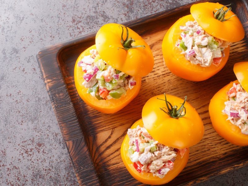 Gefüllte gelbe Tomaten mit Forellenmousse und Gemüse auf Holzbrett auf grauem Untergrund. Draufsicht.