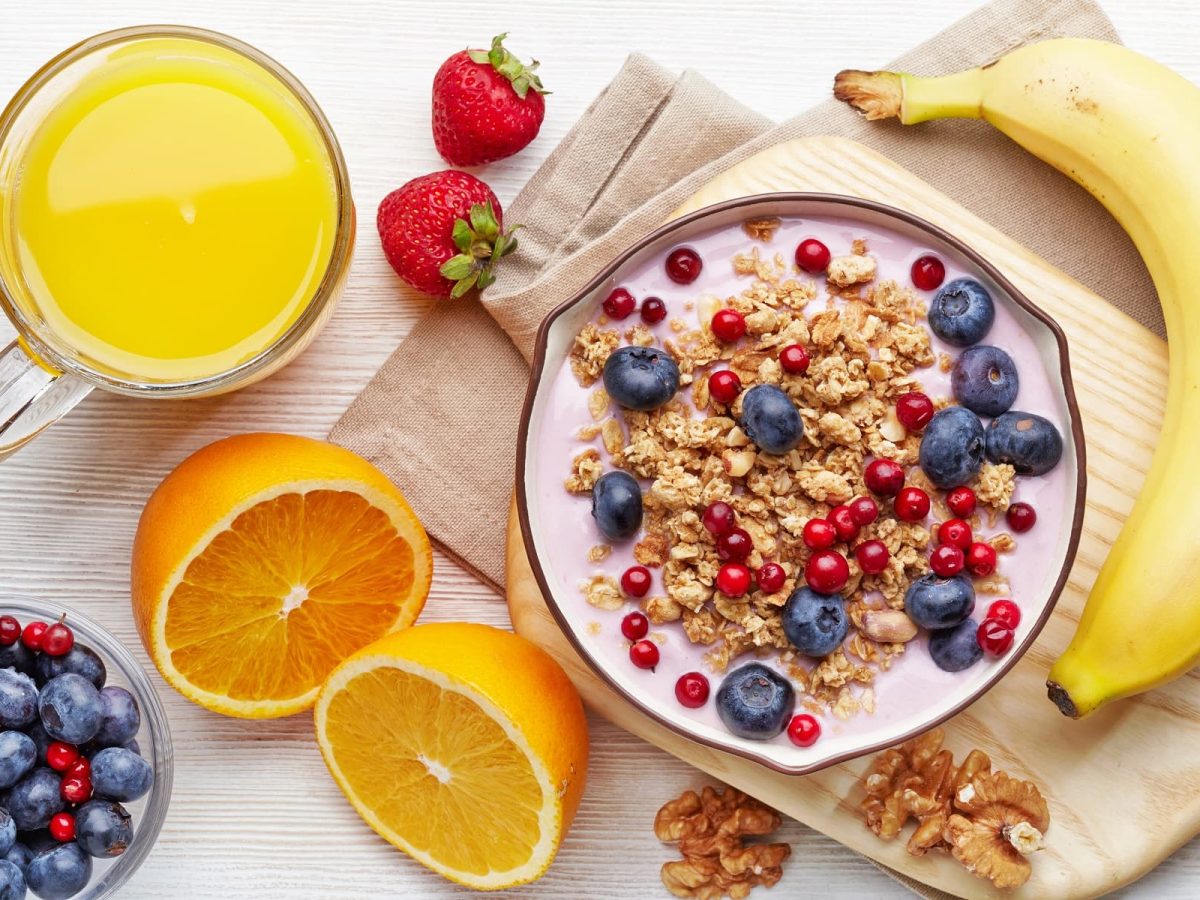 Aus der Vogelperspektive: eine Banane, zwei Orangenhälften, Beeren und einen Schale mit einem Müsli liegen auf einem weißen Tisch.