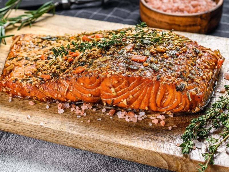 Gewürzlachs vom Grill auf Holzbrett mit Kräutern. Daneben rosa Salz. Draufsicht.