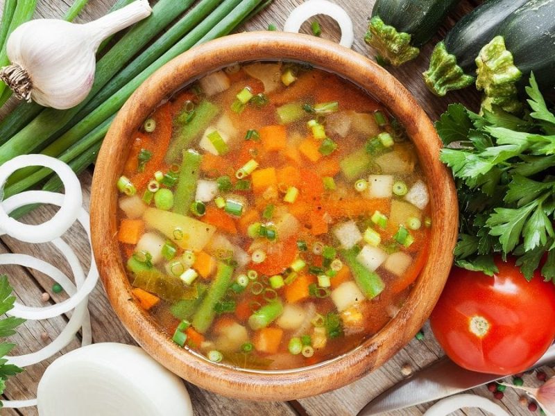 Aus der Vogelperspektive: Ein Topf grüne Bohnensuppe. Drumherum liegen Zucchini, Zwiebeln, Knoblauch und Tomaten.