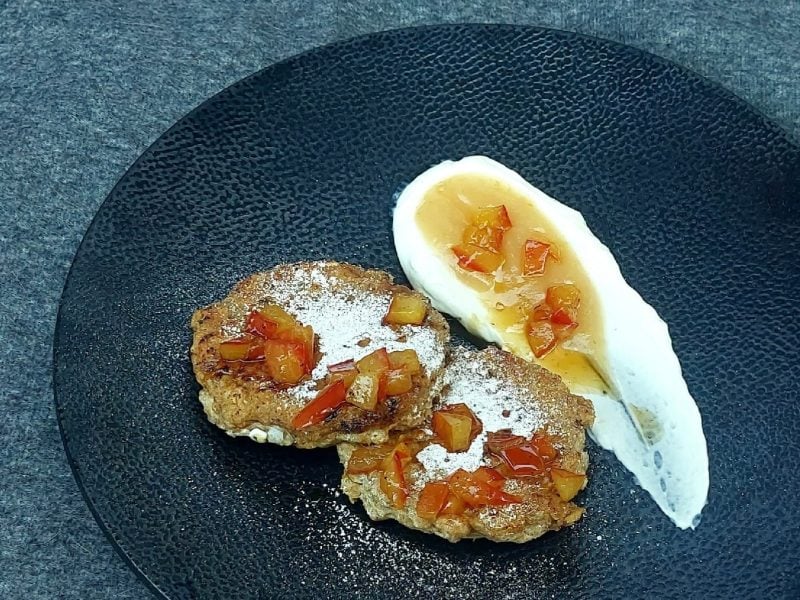 Ein schwarzer Teller mit zwei Hafer-Pancakes, Skyr , Apflemus und Apfelstückchen auf einem matt-grauen Untergrund.