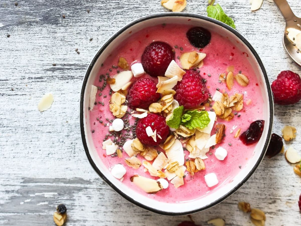 Eine Schüssel mit dem Himbeer-Kokos-Smoothie auf einem grauen Holztisch. Obendrauf frische Himbeeren, Kokoschips und Minze.