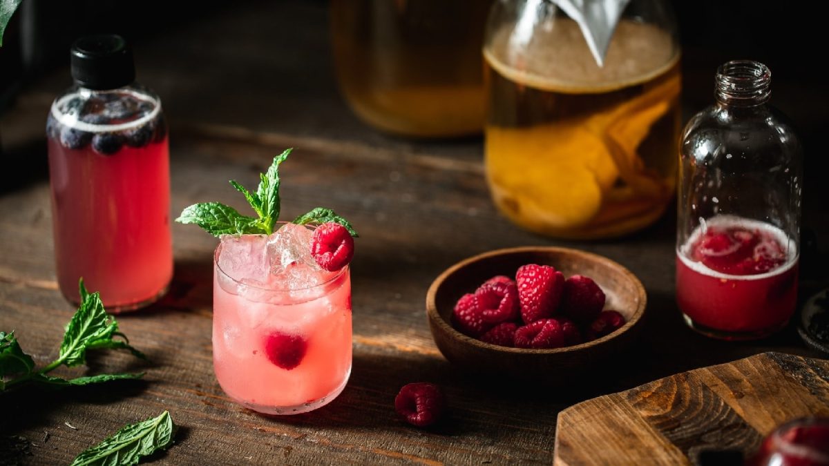 Ein Glas mit dem Himbeer-Kombucha auf einem dunklem Holzbrett mit allen weiteren Zutaten.