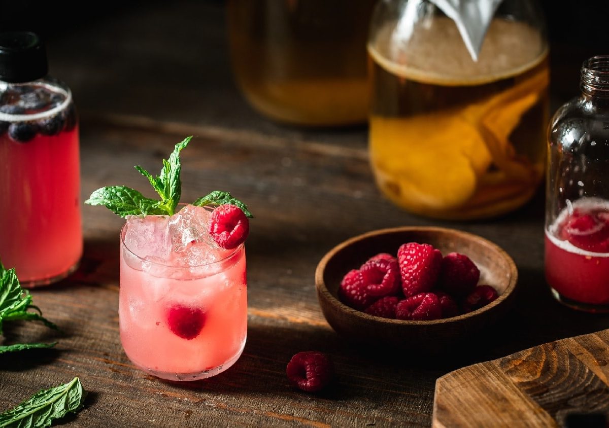 Ein Glas mit dem Himbeer-Kombucha auf einem dunklem Holzbrett mit allen weiteren Zutaten.