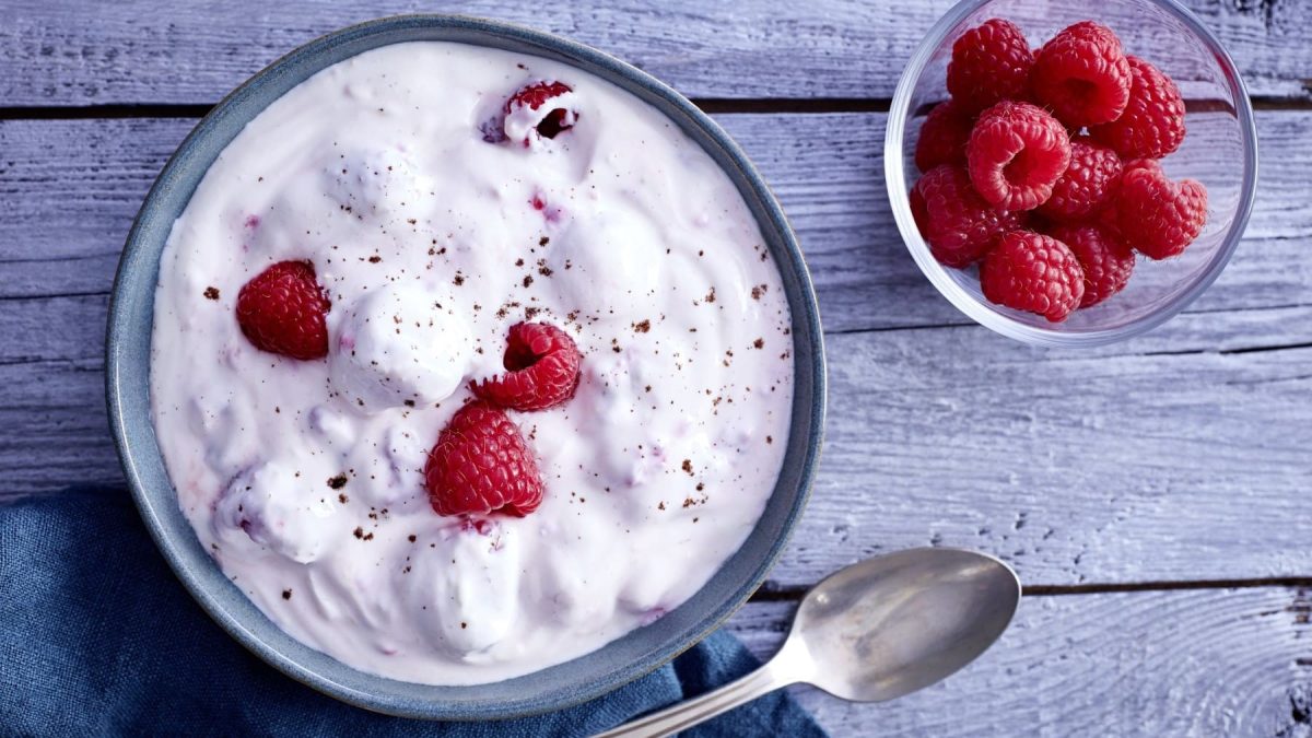 Schüssel mit Himbeer-Traum neben einem Löffel und einer kleineren Schüssel mit Himbeeren vor blauem Hintergrund.