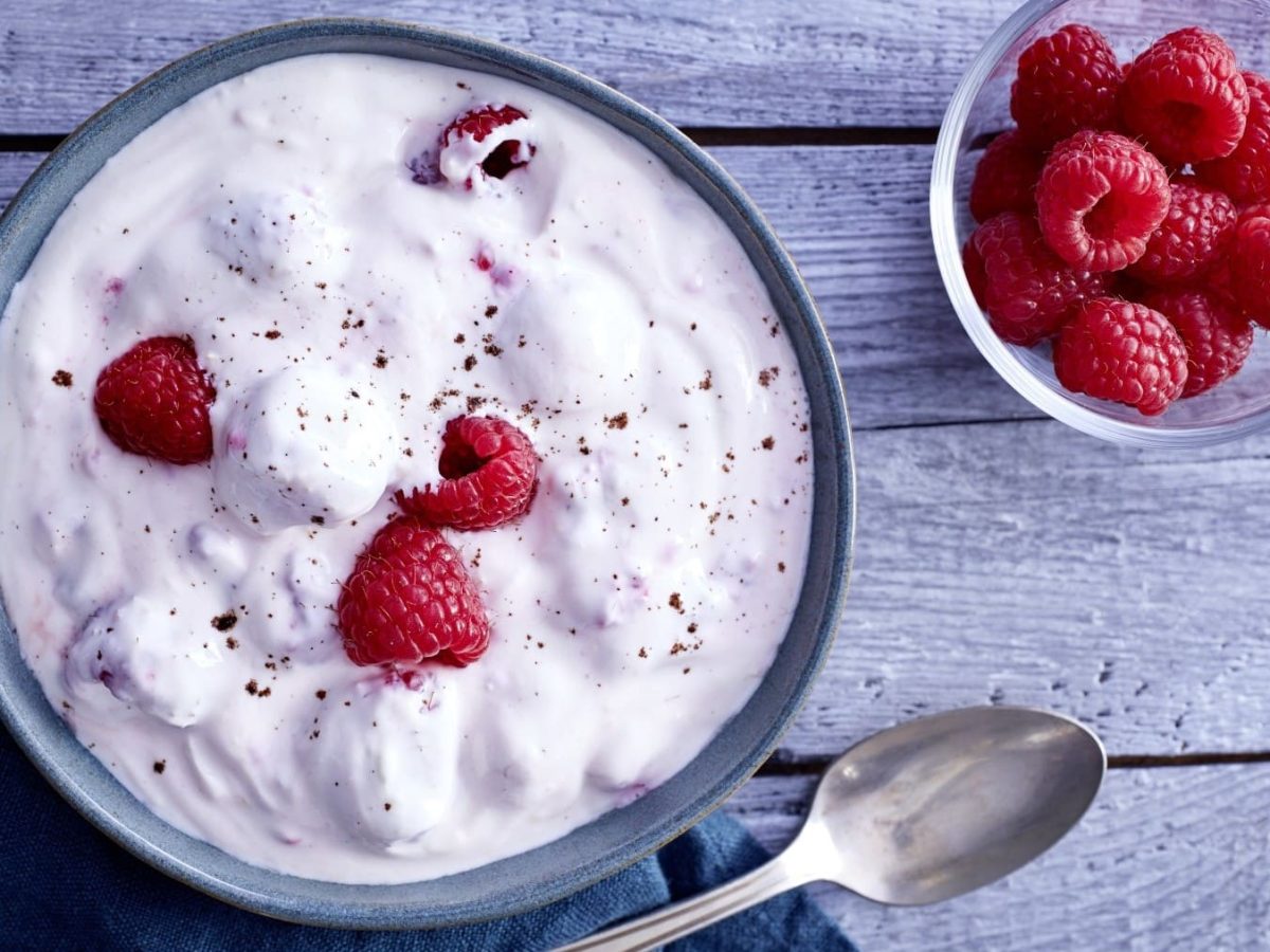 Schüssel mit Himbeer-Traum neben einem Löffel und einer kleineren Schüssel mit Himbeeren vor blauem Hintergrund.