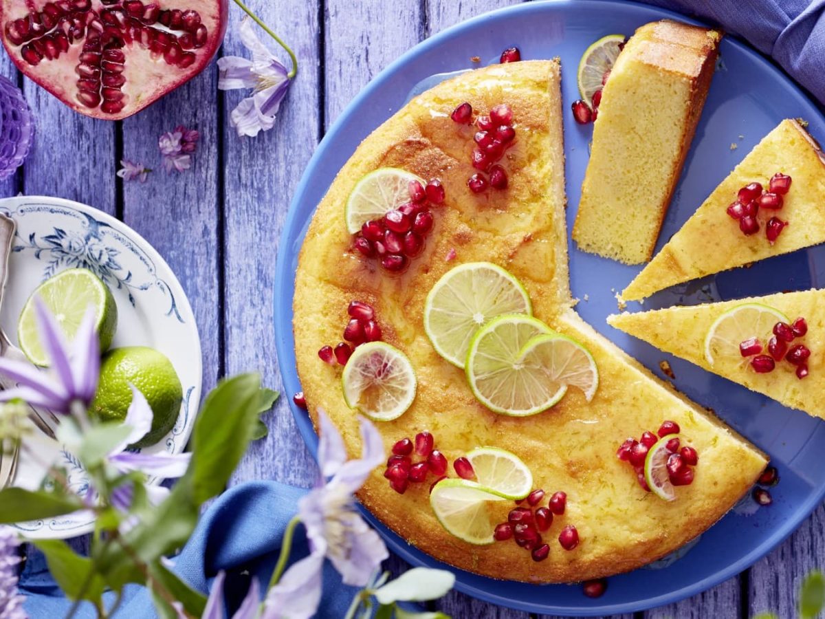 Angeschnittenet Joghurt-Limetten-Kuchen mit Granatapfelkernen und Limettenscheiben verziert auf einem blauem Teller neben Blumen und Geschirr.