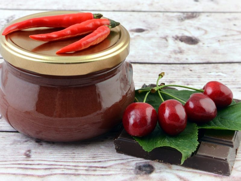 Chili-Kirschmarmelade mit Schokolade im Glas auf auf hellem Holz. Daneben Kirschen, Blätter und Schokolade. Frontalansicht.