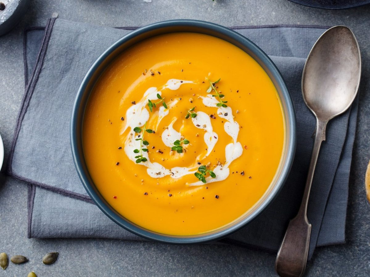 Eine Schale Kokos-Kürbissuppe in der Draufsicht, daneben ein Löffel und eine Scheibe Baguette sowie eine Schüssel mit Salz und Kernen.