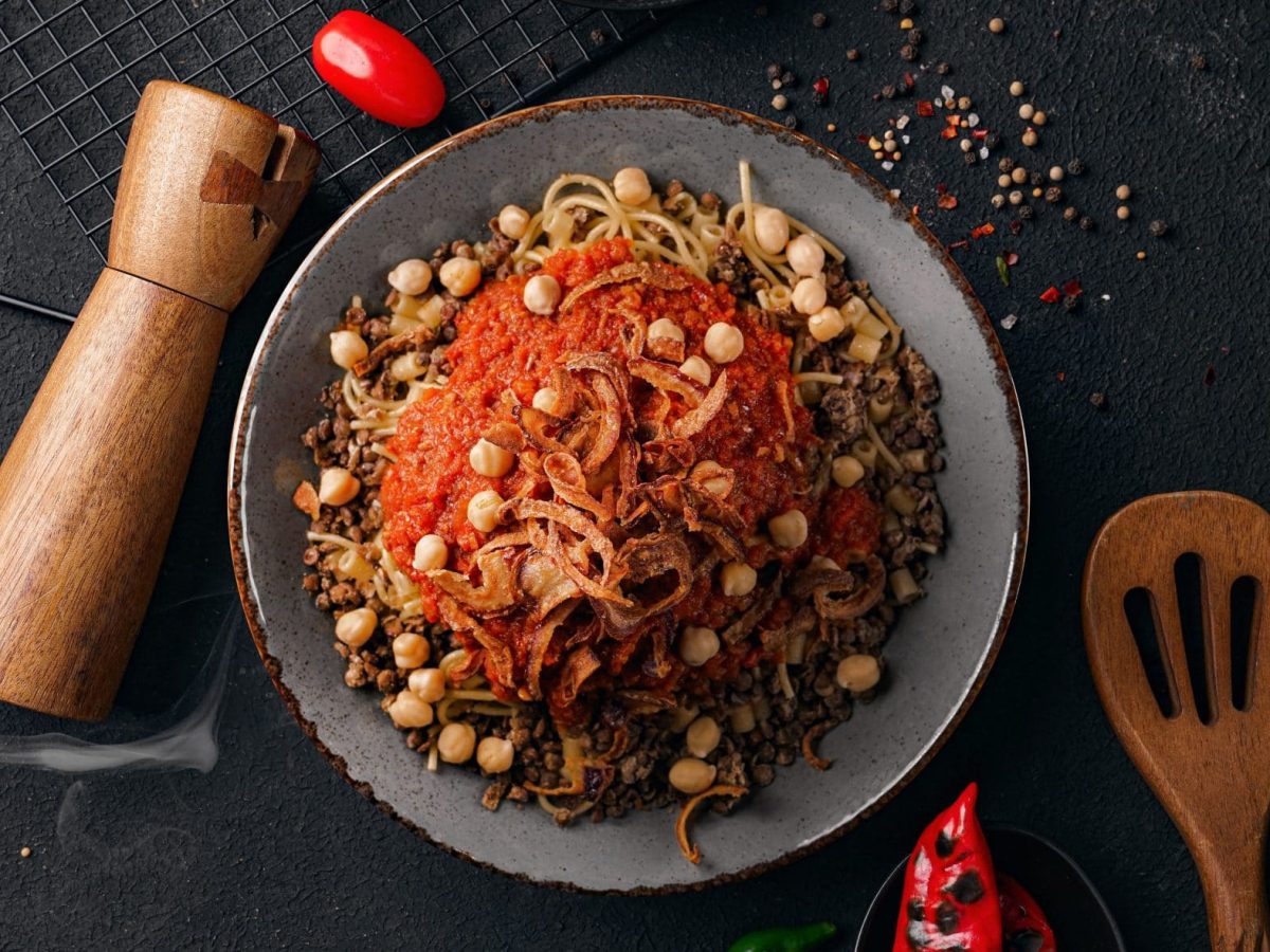 Koshari ägyptisches Streetfood auf grauem Teller und dunklem Untergrund. Danebem Pfeffermühle, Pfefferkörner und Küchenutensilien. Unten geröstete Chili. Draufsicht.