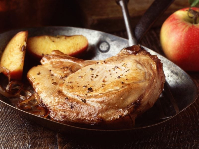 Iberico Koteletts mit Apfelweinsauce in Gusspfanne auf Holztisch. Daneben Äpfel. Frontalansicht.