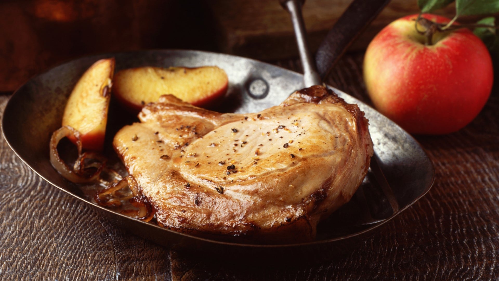 Iberico Koteletts mit Apfelweinsauce in Gusspfanne auf Holztisch. Daneben Äpfel. Frontalansicht.