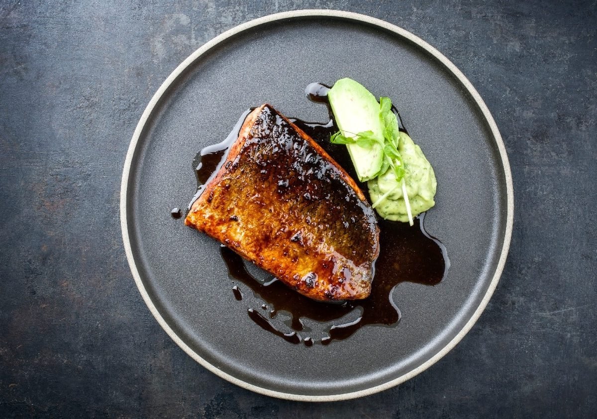 Ein schwarzer Teller mit dem Fishc und Sojalack. Rechts daneben ein Klecks mit der WAsabi-Kohlrabi-Creme und einem Stück Kohlrabi. Alles auf einem schwarzen Untergrund.