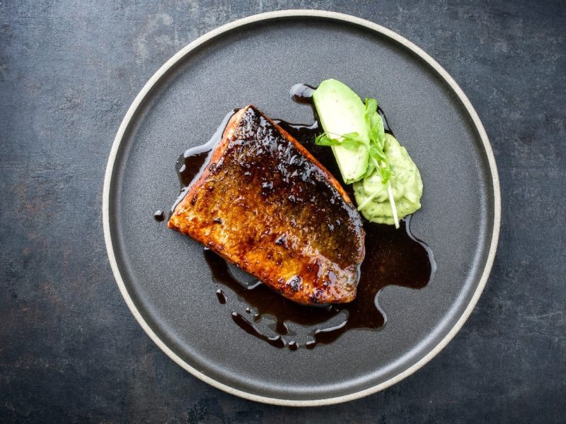 Ein schwarzer Teller mit dem Fishc und Sojalack. Rechts daneben ein Klecks mit der WAsabi-Kohlrabi-Creme und einem Stück Kohlrabi. Alles auf einem schwarzen Untergrund.